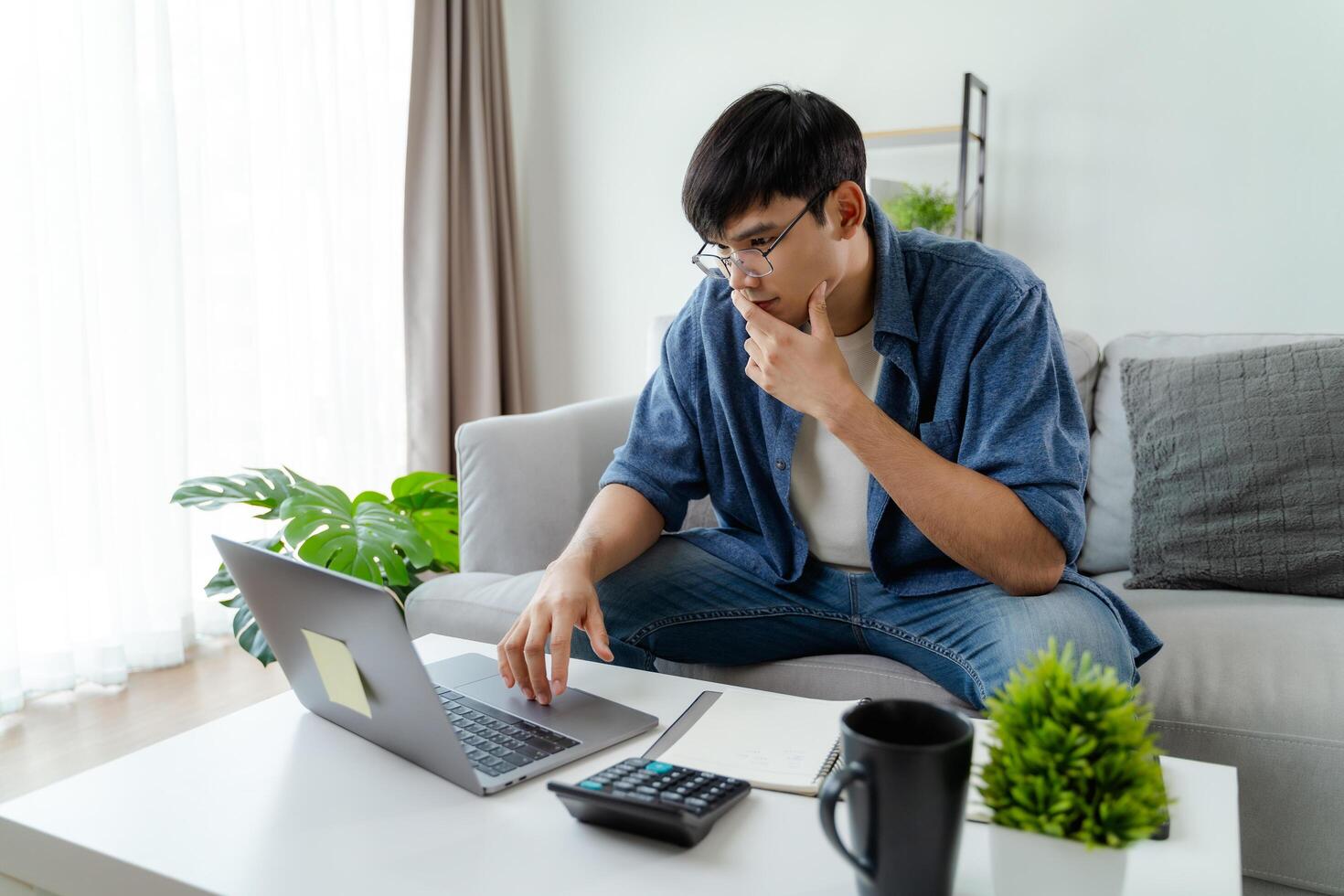 de Mens in gewoontjes kleren werken met een laptop, computer, slim telefoon, rekenmachine zittend Aan de sofa in de leven kamer Bij huis, werken van huis concept. foto