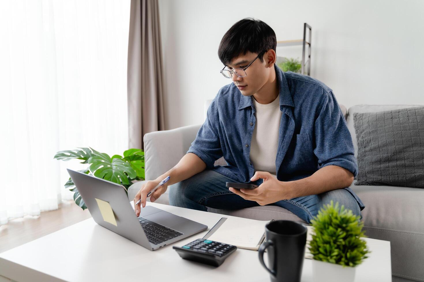 de Mens in gewoontjes kleren werken met een laptop, computer, slim telefoon, rekenmachine zittend Aan de sofa in de leven kamer Bij huis, werken van huis concept. foto