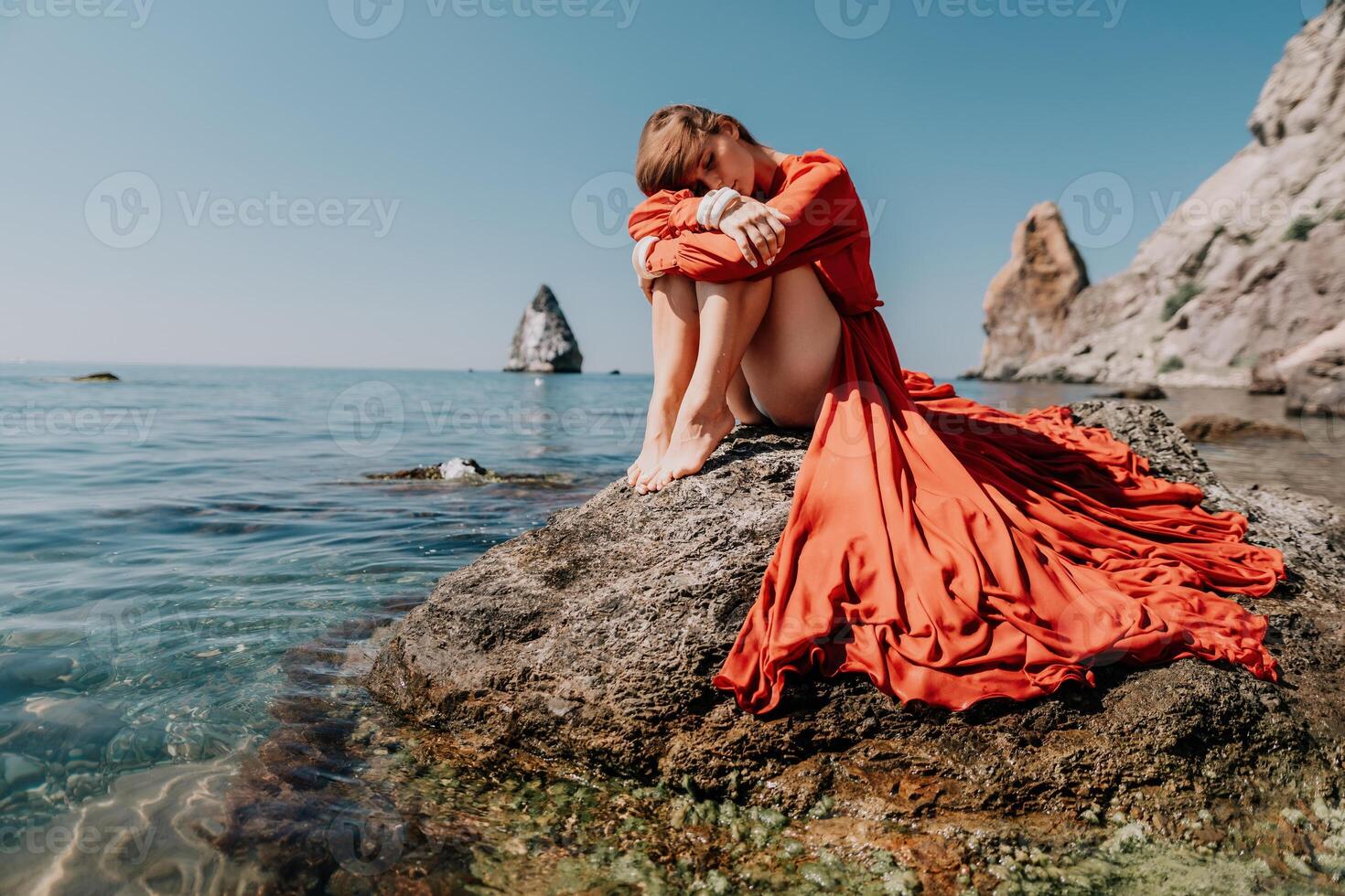 vrouw reizen zee. gelukkig toerist in rood jurk genieten nemen afbeelding buitenshuis voor herinneringen. vrouw reiziger poseren Aan de rots Bij zee baai omringd door vulkanisch bergen, sharing reizen avontuur reis foto