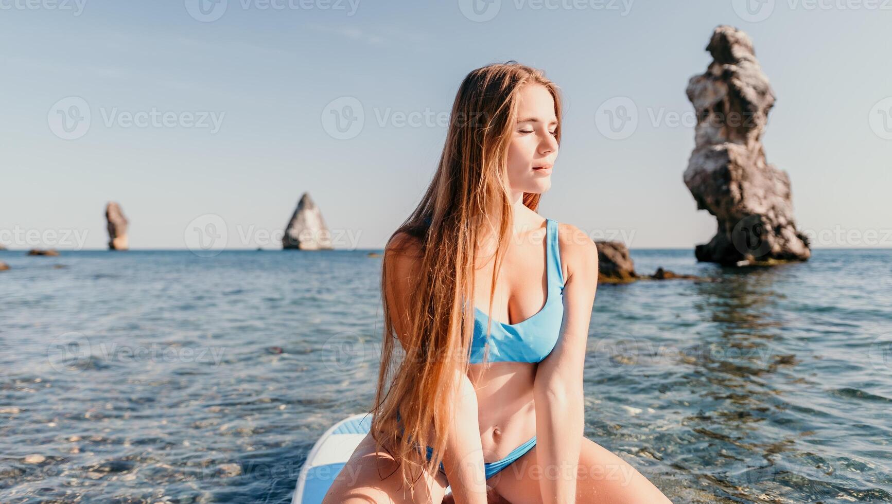 vrouw zee sup. dichtbij omhoog portret van gelukkig jong Kaukasisch vrouw met lang haar- op zoek Bij camera en lachend. schattig vrouw portret in een blauw bikini poseren Aan sup bord in de zee foto