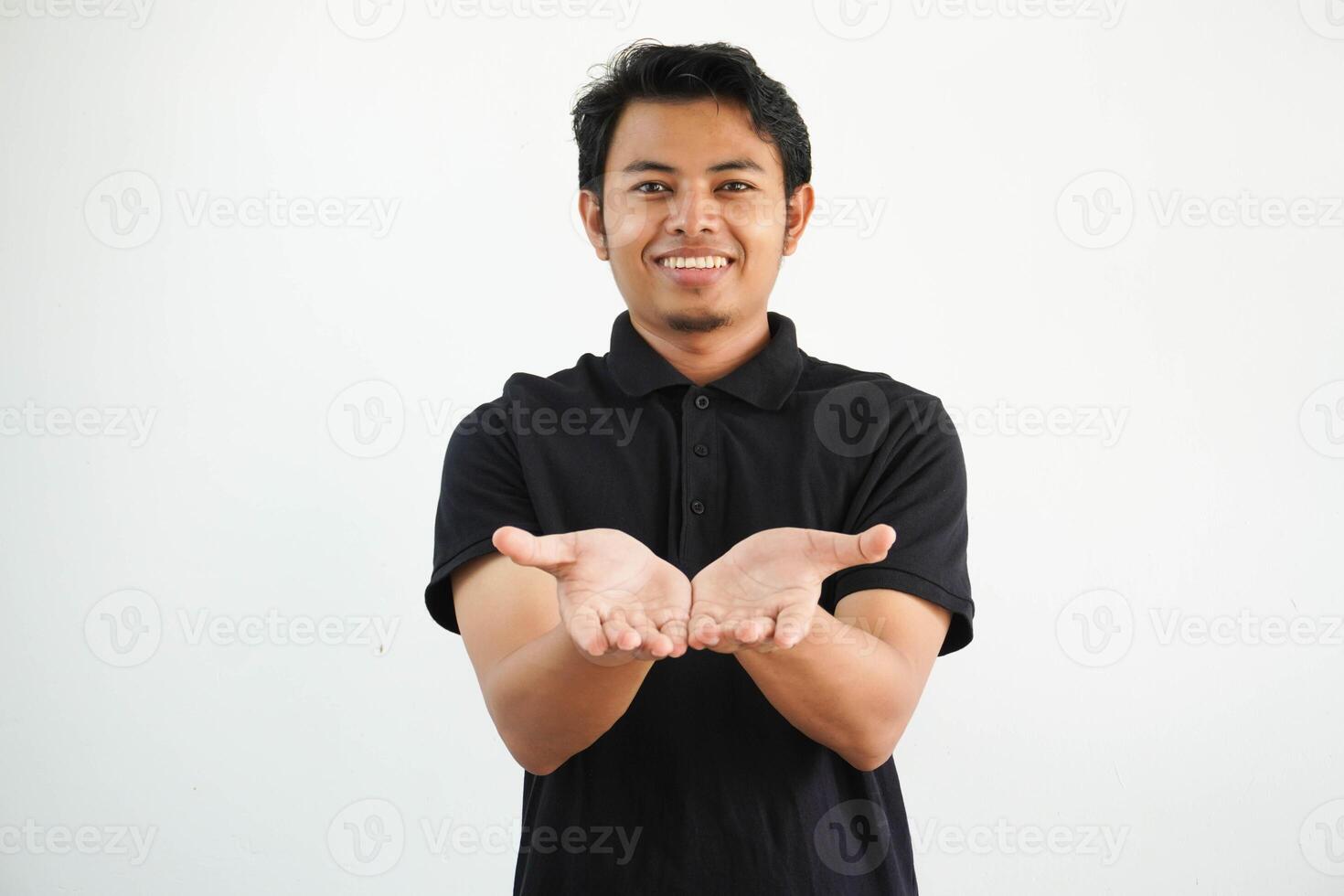 glimlachen jong Aziatisch Mens poseren Aan een wit backdrop Holding iets met handpalmen, aanbieden naar camera, vervelend zwart polo t shirt. foto