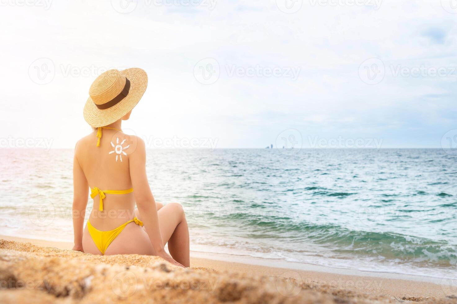 een jong Kaukasisch vrouw in een zwempak en een rietje hoed zit Aan de strand in voorkant van de zee met haar terug in de kader. de zon Aan een vrouw terug is getrokken met zonnescherm foto