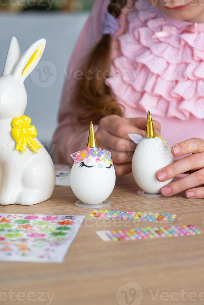 een schattig meisje met roze konijn oren maakt een Pasen ambacht - siert een ei in de het formulier van een eenhoorn met strass steentjes, Hoorn, bloemen in de interieur van een huis met planten. foto