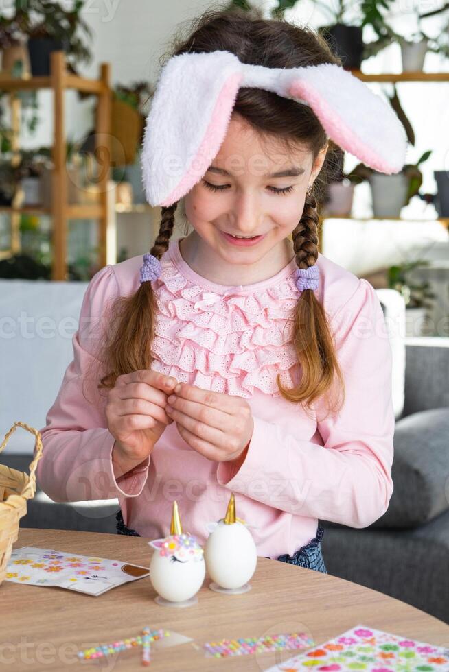 een schattig meisje met roze konijn oren maakt een Pasen ambacht - siert een ei in de het formulier van een eenhoorn met strass steentjes, Hoorn, bloemen in de interieur van een huis met planten. foto