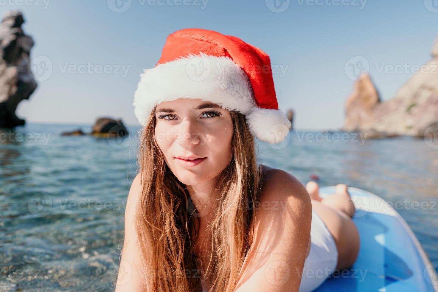 vrouw zee sup. dichtbij omhoog portret van gelukkig jong Kaukasisch vrouw met lang haar- in de kerstman hoed op zoek Bij camera en lachend. schattig vrouw portret in een wit bikini poseren Aan sup bord in de zee foto