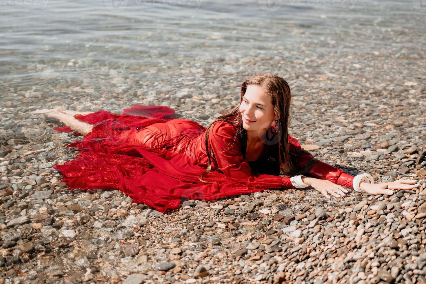 vrouw reizen zee. gelukkig toerist in rood jurk genieten nemen afbeelding buitenshuis voor herinneringen. vrouw reiziger poseren in zee strand, omringd door vulkanisch bergen, sharing reizen avontuur reis foto