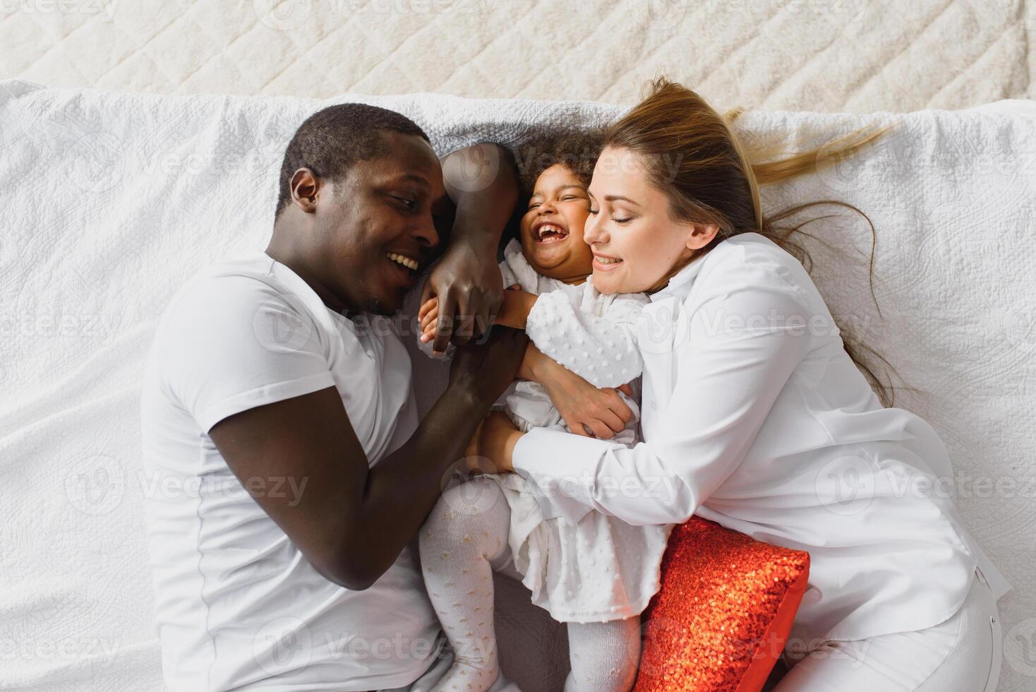 portret van gelukkig multiraciaal jong familie aan het liegen Aan knus wit bed Bij huis, glimlachen Internationale mam en vader ontspannende met weinig biraciaal meisje kind poseren voor afbeelding in slaapkamer foto