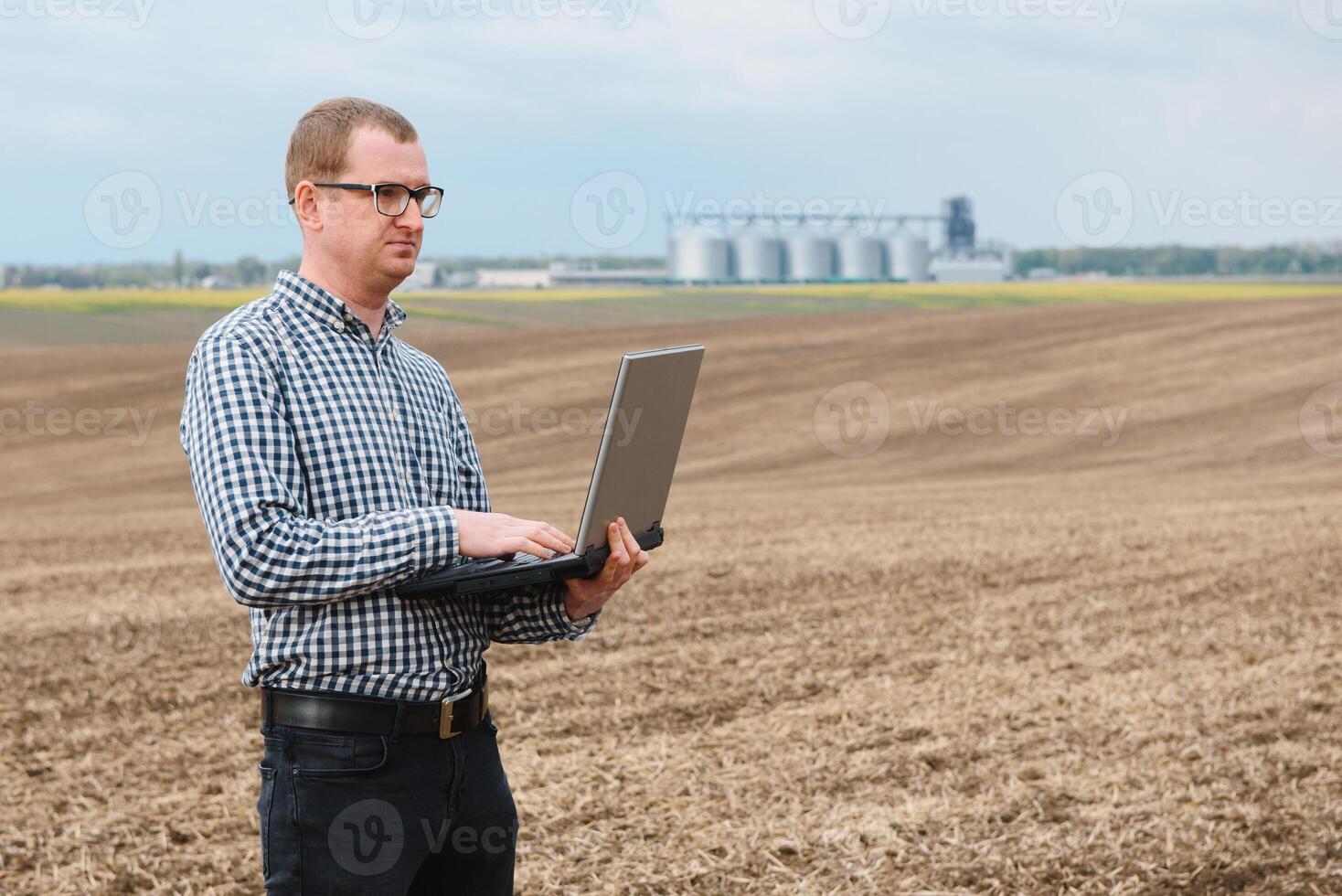 modern boer controle zijn veld- fabriek en werken Aan laptop computer tegen maïs droger silo's in concept van industrieel en landbouw foto