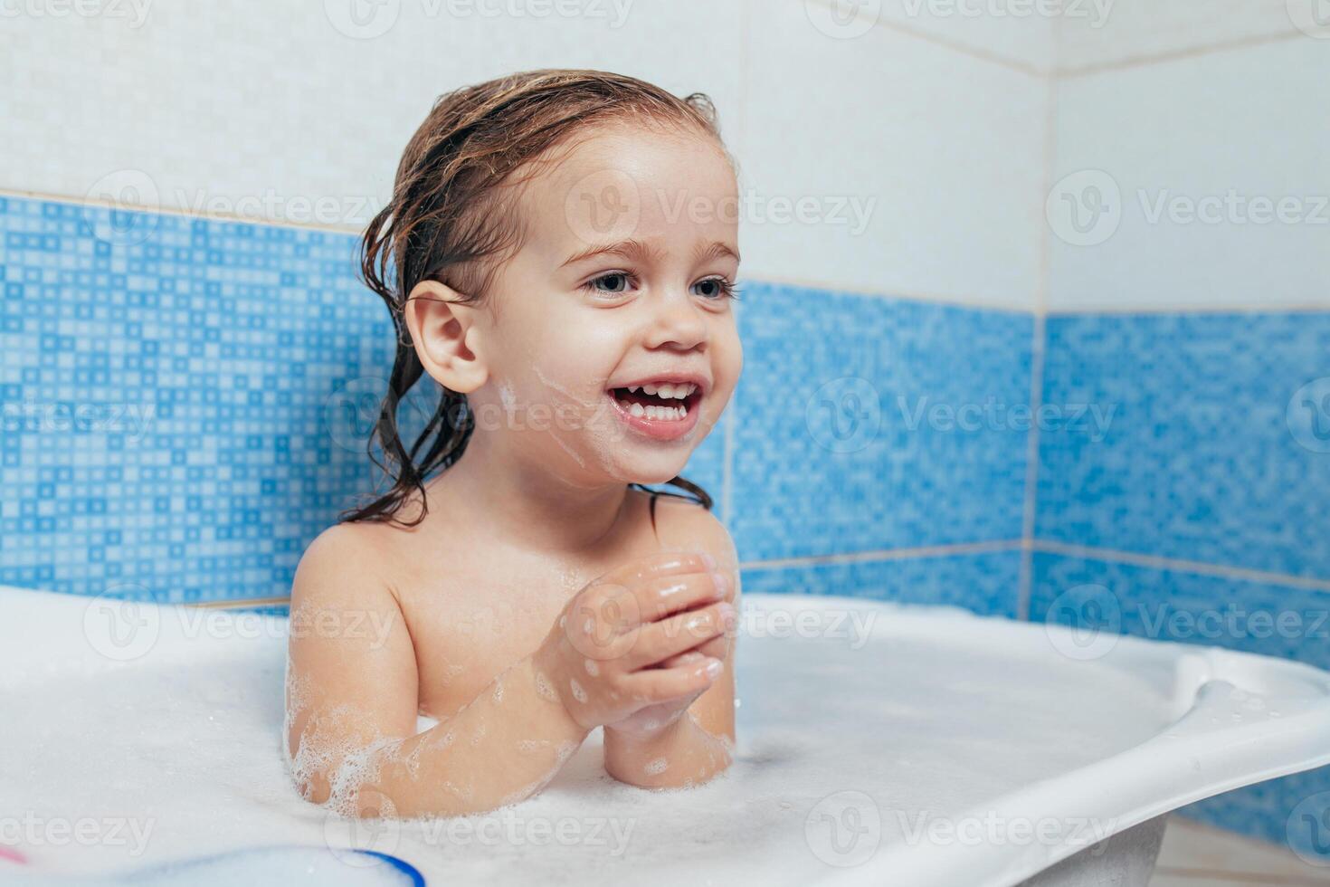 pret vrolijk gelukkig kleuter baby nemen een bad spelen met schuim bubbels. weinig kind in een bad. glimlachen kind in badkamer Aan blauw achtergrond. hygiëne en Gezondheid zorg. foto