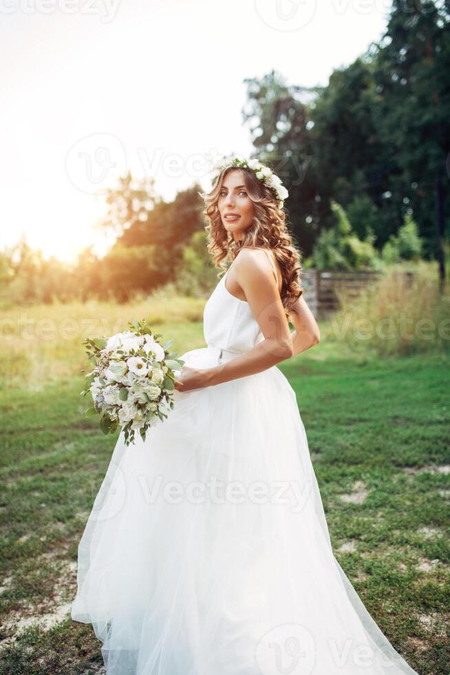 een schattig gekruld vrouw in een wit bruiloft jurk met een bruiloft boeket en krans in haar haar- staand terug naar de camera in natuur. concept ontsnapte bruid. vooruit naar een gelukkig helder toekomst ren weg foto