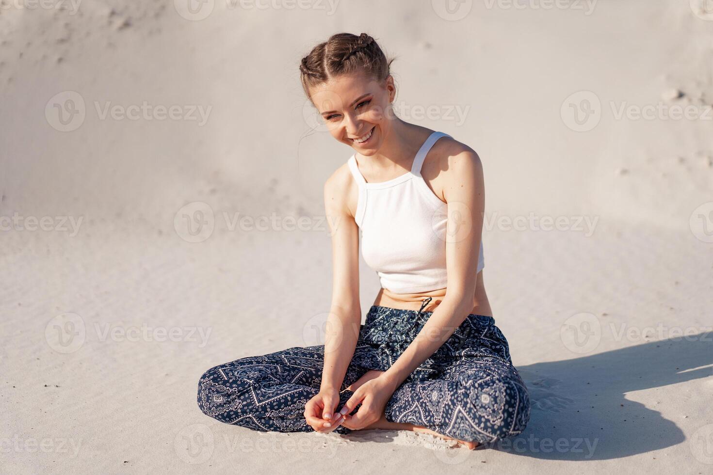 een mooi jong Kaukasisch meisje in een wit top en breed broek zit in een lotus positie Aan de strand Aan de zand. foto