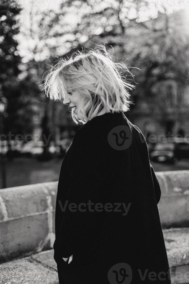 zorgeloos blond vrouw glimlachen in zwart jas, wandelen door park. blond vrouw in zwart jas wandelen gelukkig in park, uitstralend vreugde en zorgeloos geest foto