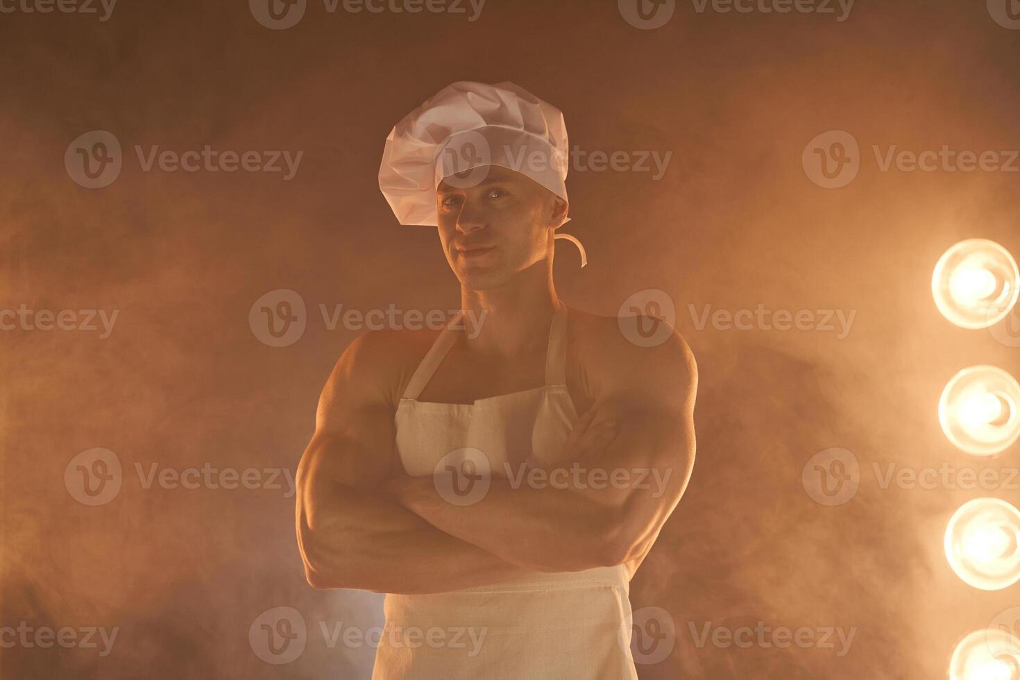 portret van gespierd chef vervelend wit schort en chef hoed, poseren met gevouwen armen Aan rokerig achtergrond foto