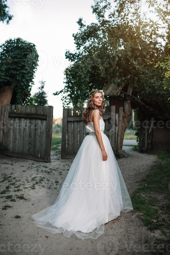 een schattig gekruld vrouw in een wit bruiloft jurk met een bruiloft boeket en krans in haar haar- staand terug naar de camera in natuur. concept ontsnapte bruid. vooruit naar een gelukkig helder toekomst ren weg foto