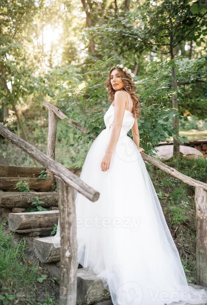een schattig gekruld vrouw in een wit bruiloft jurk met een bruiloft boeket en krans in haar haar- staand terug naar de camera in natuur. concept ontsnapte bruid. vooruit naar een gelukkig helder toekomst ren weg foto