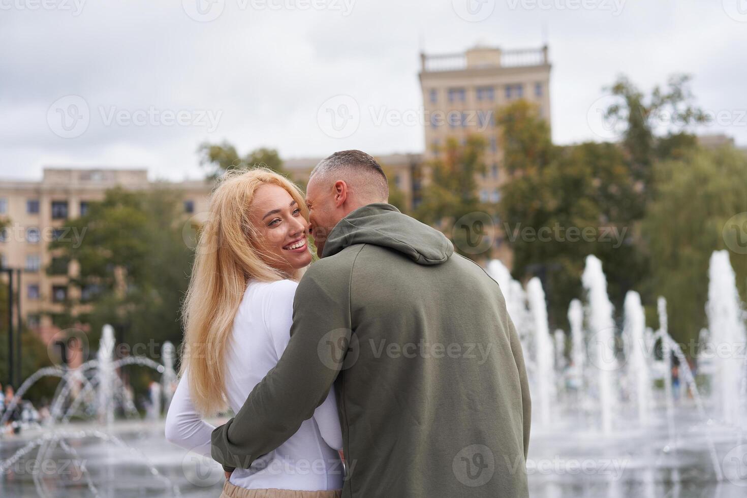 romantisch wandelen. jong paar in liefde omarmen in de buurt fontein in stedelijk park, Mens fluisteren complimenten naar mooi vriendin foto