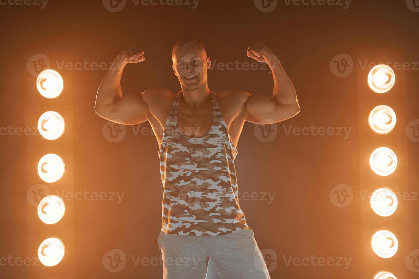 actief levensstijl. professioneel bodybuilder tonen biceps en glimlachen Bij camera, lampen verlichting Aan achtergrond foto