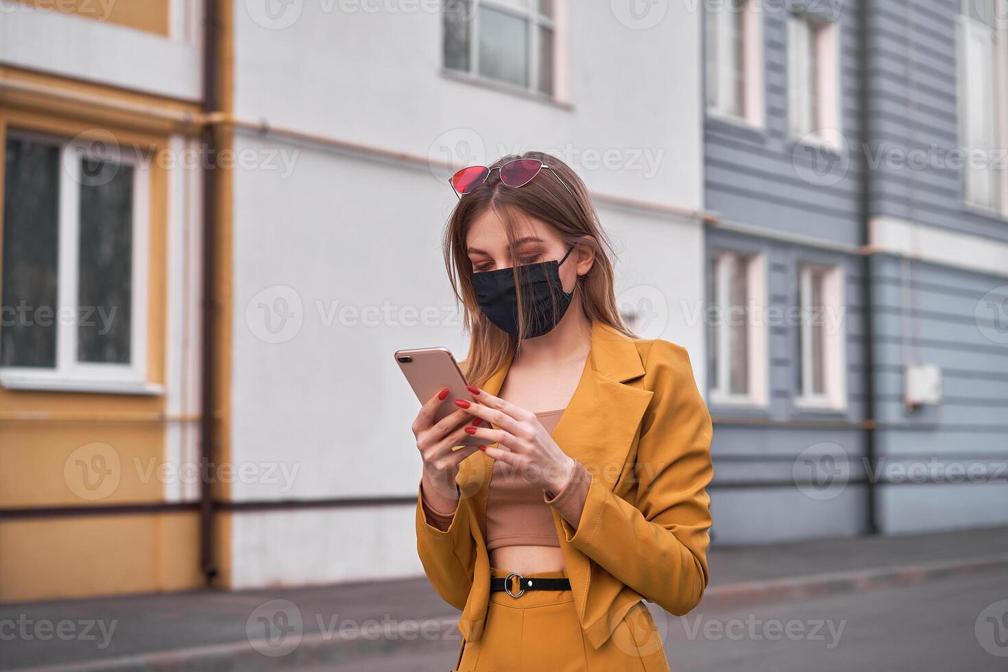 jong mooi Kaukasisch meisje 20 jaren slijtage zwart gezicht masker bescherming tegen epidemie coronavirus covid-19 wandelingen naar beneden de straat looks Bij de telefoon scherm. foto