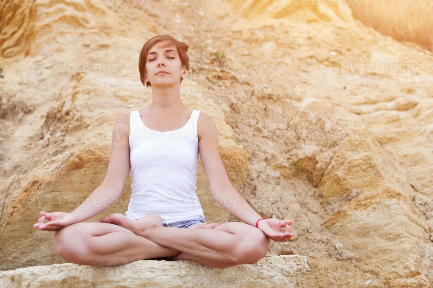 een mooi jong meisje met kort haar- is gekleed in shorts en een wit Jersey is beoefenen yoga Aan de achtergrond van rotsen. houding van de lotus. de concept van kalmte en concentratie foto