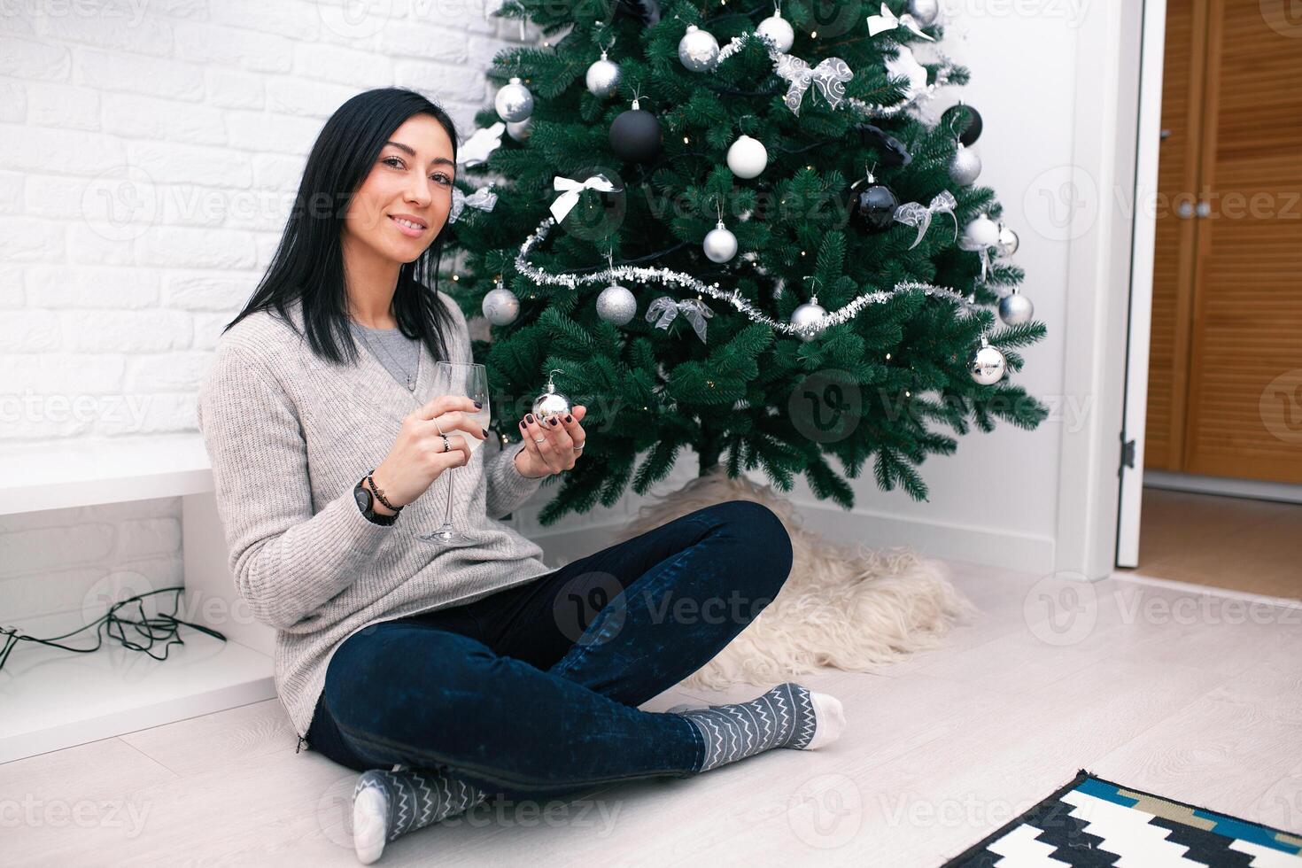 een jong vrouw zit in de buurt een Kerstmis boom, Holding een glas en een Kerstmis speelgoed. voorbereiding voor de nieuw jaar viering foto