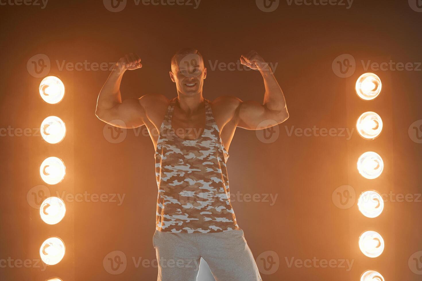 actief levensstijl. professioneel bodybuilder tonen biceps en glimlachen Bij camera, lampen verlichting Aan achtergrond foto