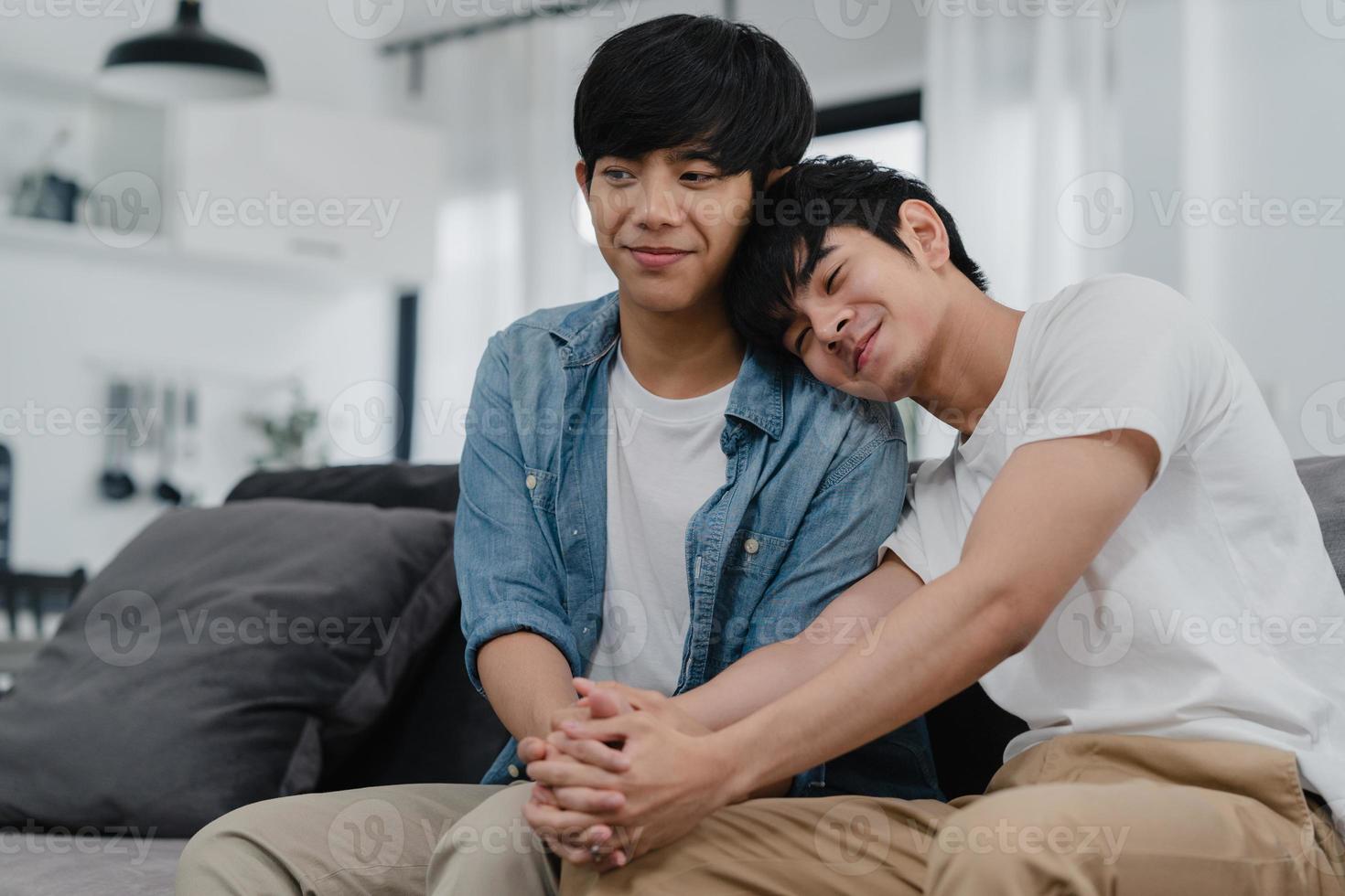 portret jong aziatisch homopaar dat zich gelukkig thuis glimlacht. Aziatische lgbtq-mannen ontspannen een brede glimlach terwijl ze naar de camera kijken terwijl ze 's ochtends op de bank in de woonkamer thuis liggen. foto