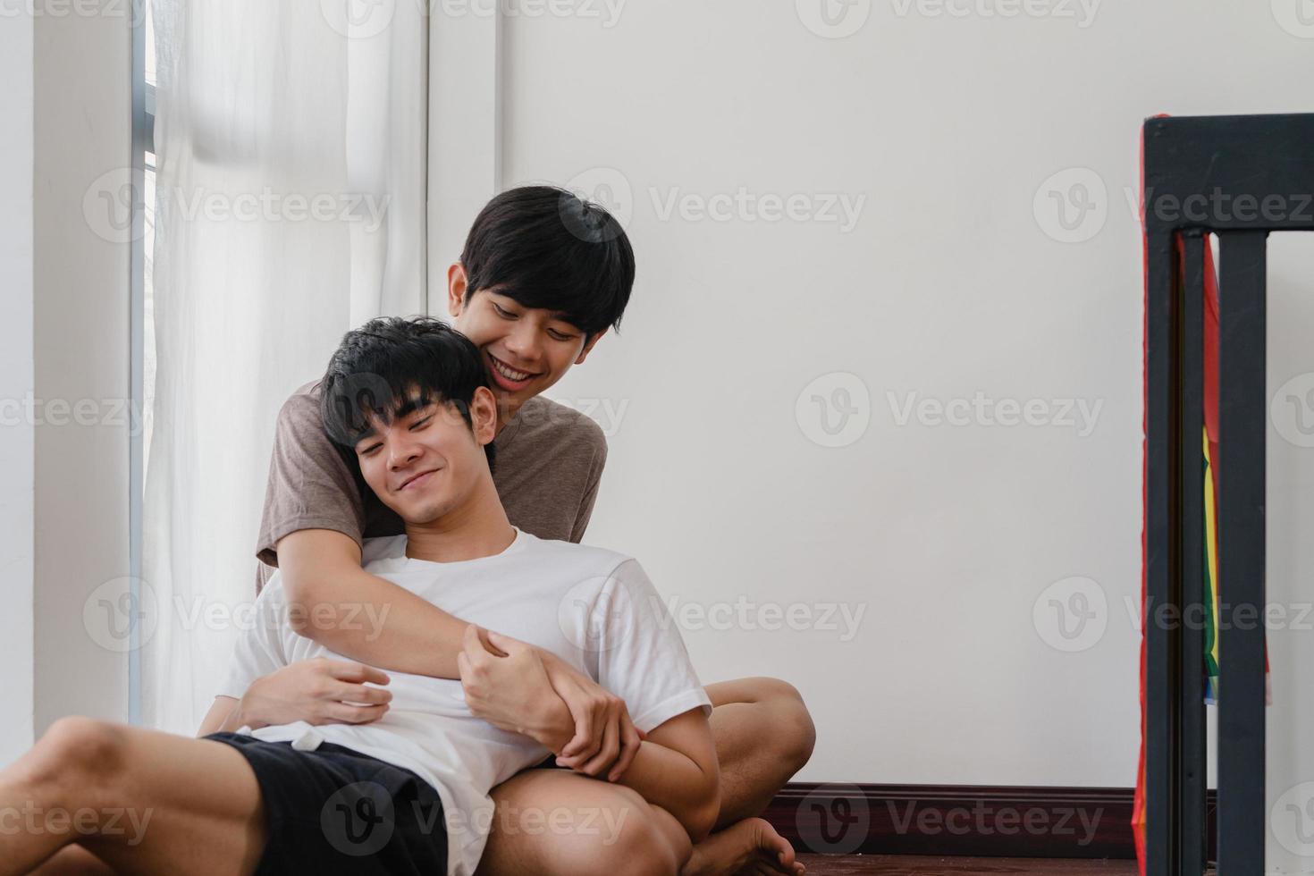 Aziatische homo paar liegen en knuffelen op de vloer thuis. jonge aziatische lgbtq-mannen zoenen gelukkig ontspannen rust samen brengen romantische tijd door in de woonkamer met regenboogvlag in het moderne huis in de ochtend. foto