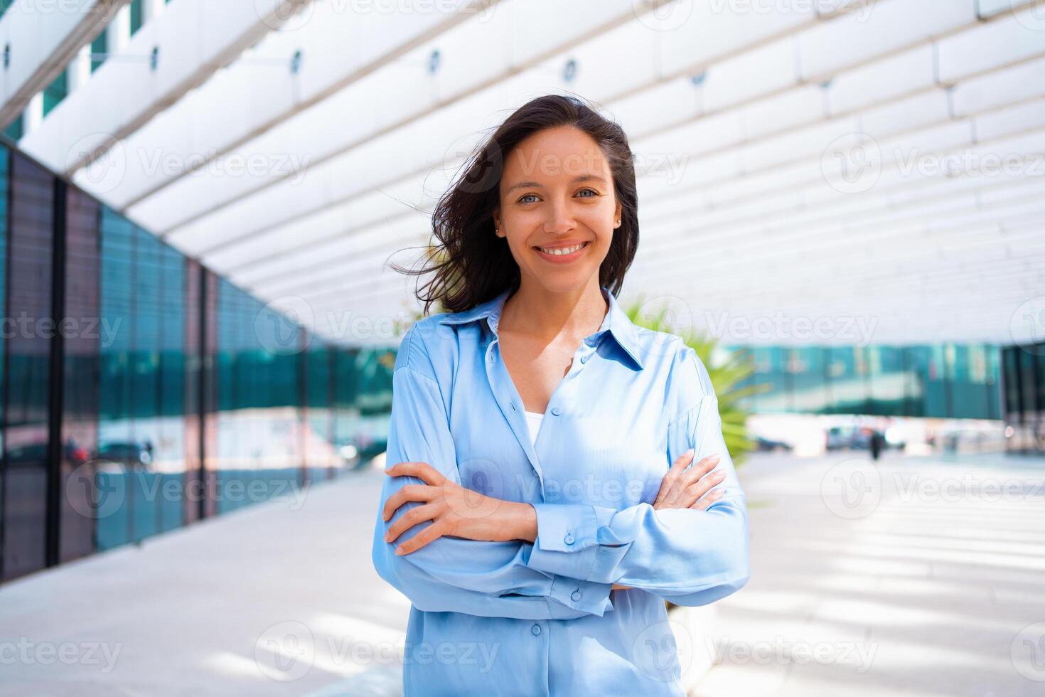 vertrouwen zakenvrouw portret met gekruiste handen. mooi bedrijf vrouw 30 jaren oud staand in de buurt kantoor gebouw gekleed blauw shirt. foto