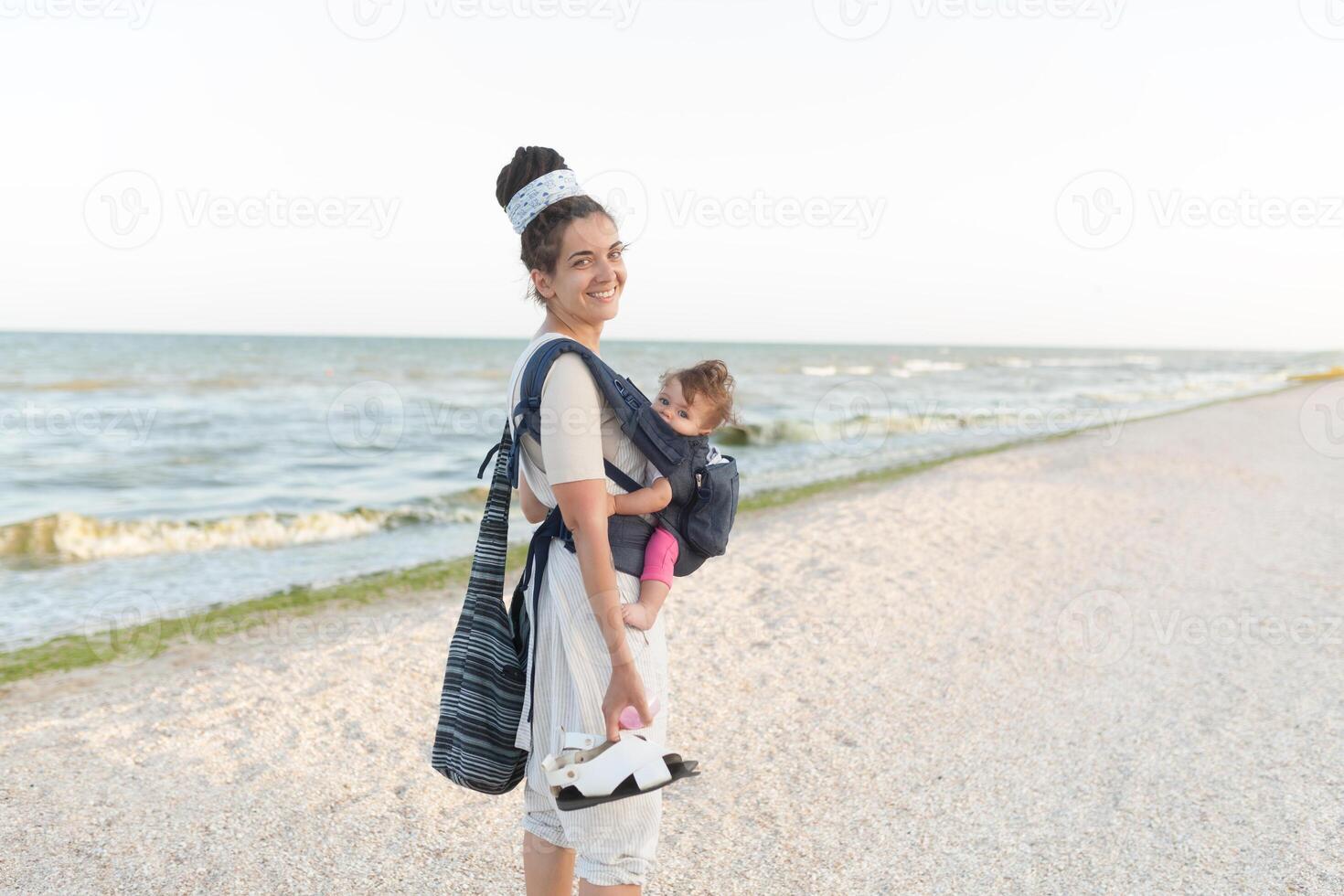 een klein kind zit in een rugzak en wandelingen langs met de moeder langs de kust. zomer familie vakantie concept foto