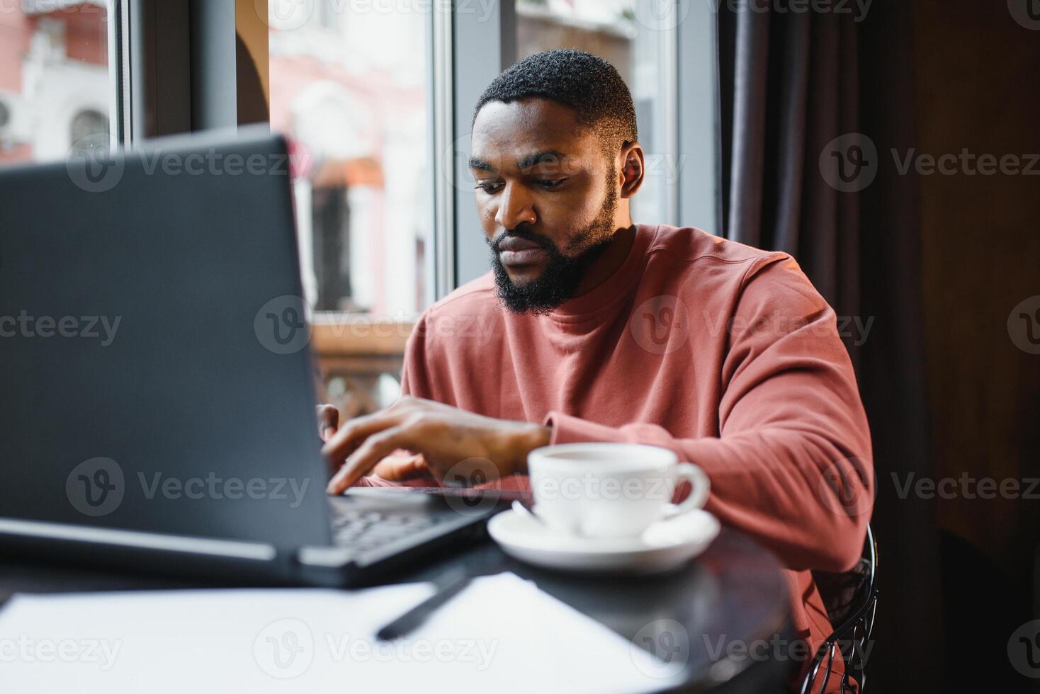 profiel portret van jong Afrikaanse online winkel manager, pratend Aan mobiel telefoon naar cliënt, overleg plegen hem Aan levering termen, zittend Bij cafe en gebruik makend van laptop computer voor werk, hebben koffie en taart. foto