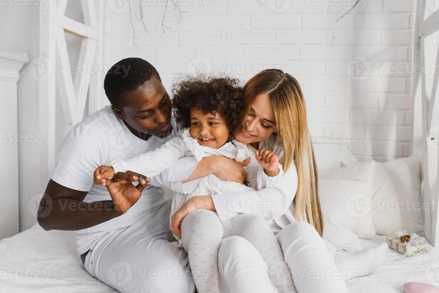 portret van gelukkig multiraciaal jong familie aan het liegen Aan knus wit bed Bij huis, glimlachen Internationale mam en vader ontspannende met weinig biraciaal meisje kind poseren voor afbeelding in slaapkamer foto