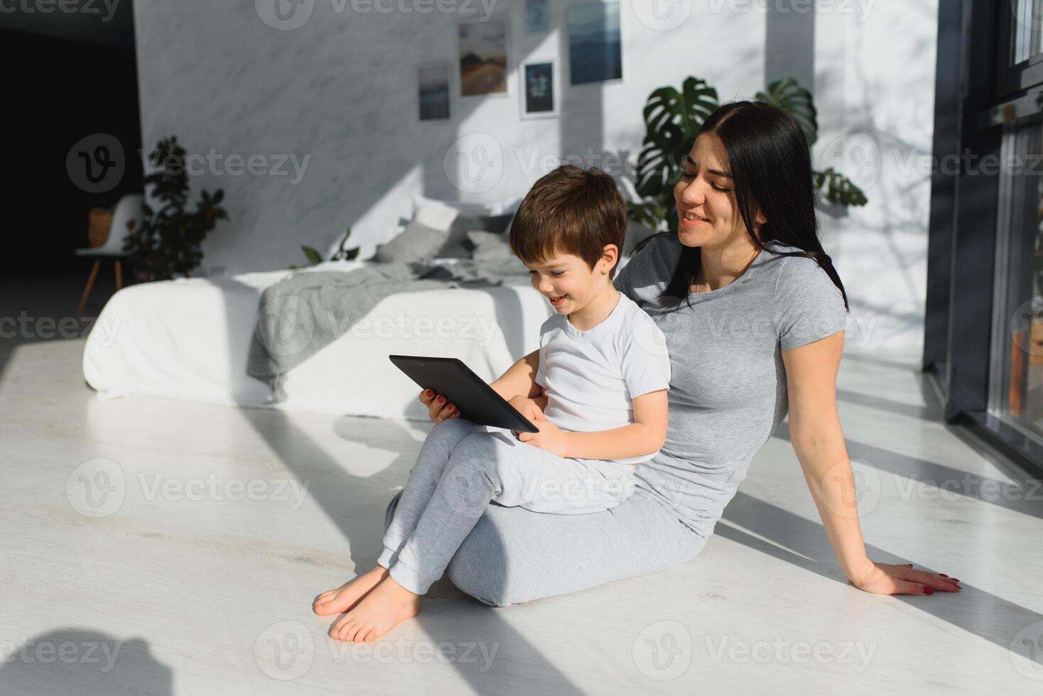 schattig weinig jongen en zijn jong moeder gebruik makend van een digitaal tablet en glimlachen Aan de verdieping Bij huis. gelukkig familie met apparaatje foto