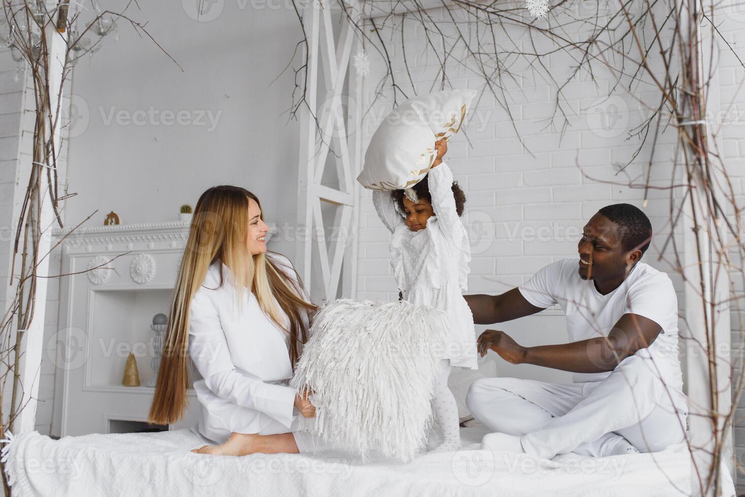 portret van gelukkig multiraciaal jong familie aan het liegen Aan knus wit bed Bij huis, glimlachen Internationale mam en vader ontspannende met weinig biraciaal meisje kind poseren voor afbeelding in slaapkamer foto