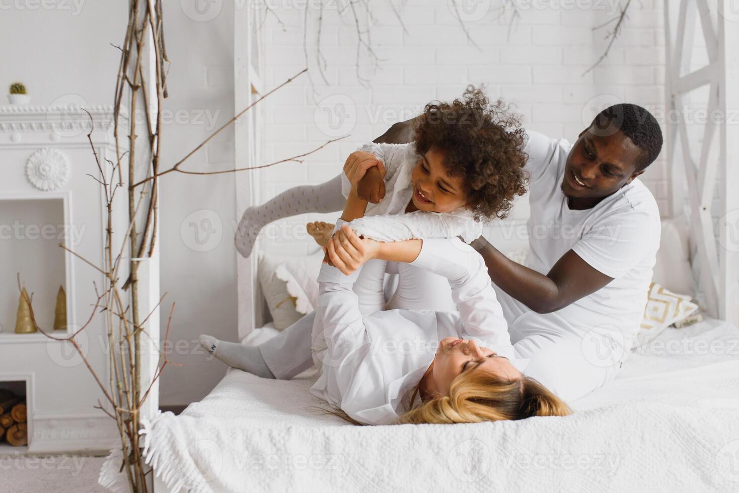 portret van gelukkig multiraciaal jong familie aan het liegen Aan knus wit bed Bij huis, glimlachen Internationale mam en vader ontspannende met weinig biraciaal meisje kind poseren voor afbeelding in slaapkamer foto