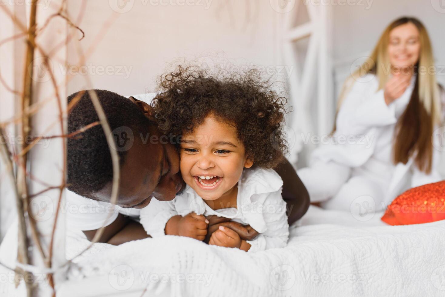 portret van gelukkig multiraciaal jong familie aan het liegen Aan knus wit bed Bij huis, glimlachen Internationale mam en vader ontspannende met weinig biraciaal meisje kind poseren voor afbeelding in slaapkamer foto