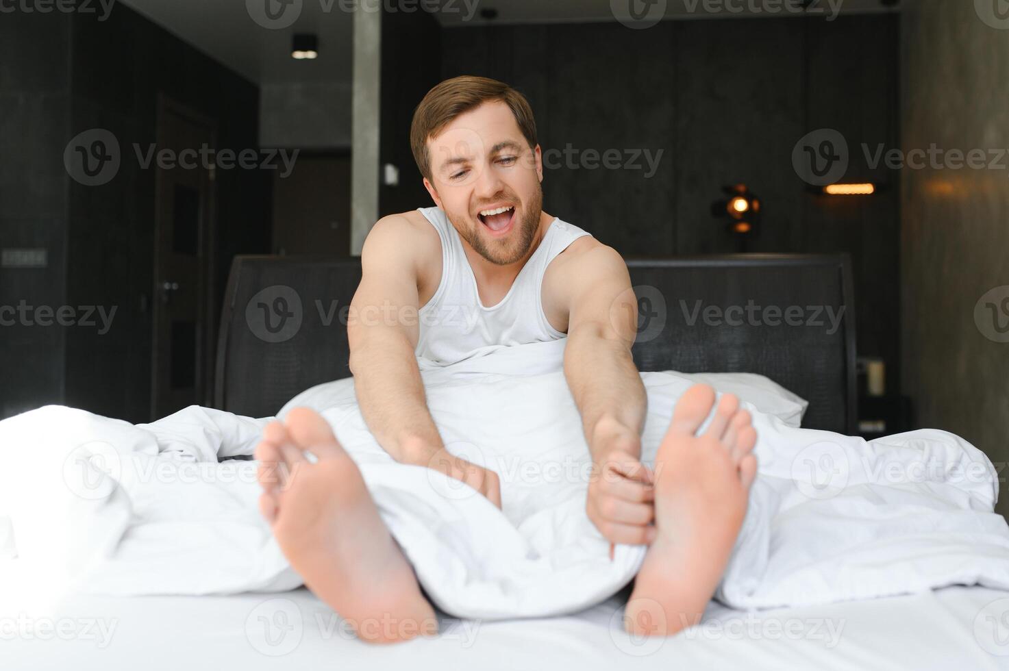 jong Mens ontwaken omhoog en stijgen handen uitrekken Aan wit bed in de ochtend- met zonlicht door venster foto