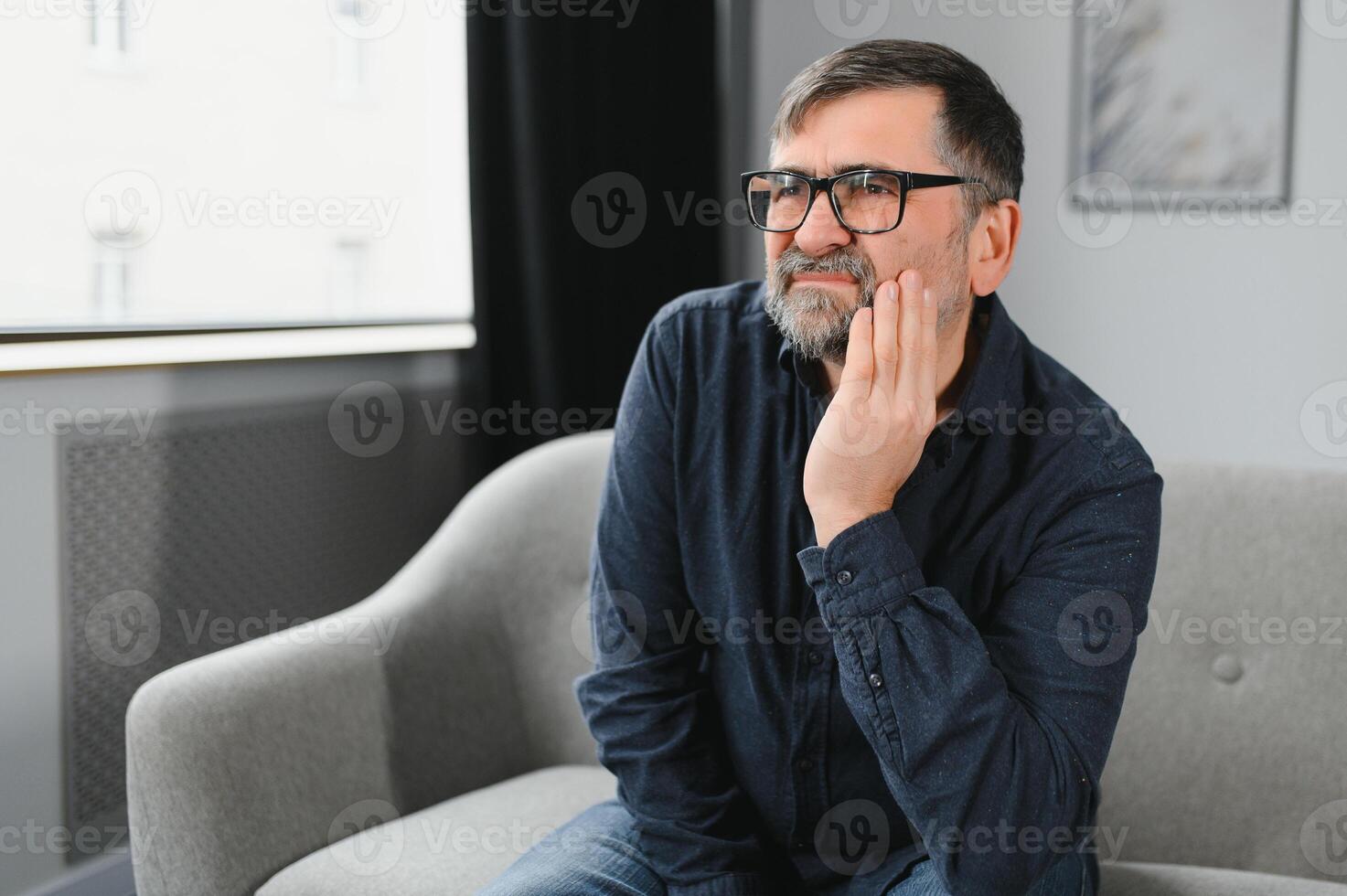 tandheelkundig zorg, tand gevoeligheid concept. portret van ongelukkig volwassen Mens lijden van vreselijk kiespijn, aanraken wang gevoel acuut pijn, oud mannetje zittend Aan de bankstel Bij huis, vrij kopiëren ruimte foto