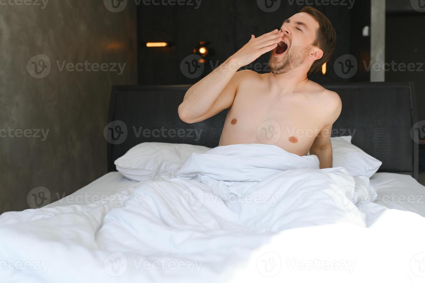 jong Mens ontwaken omhoog en stijgen handen uitrekken Aan wit bed in de ochtend- met zonlicht door venster foto