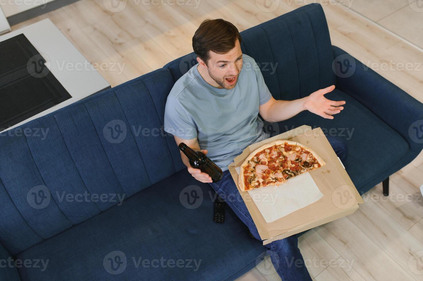 gelukkig jong Mens drinken bier en aan het eten pizza wanneer aan het kijken spel Aan TV Bij huis foto