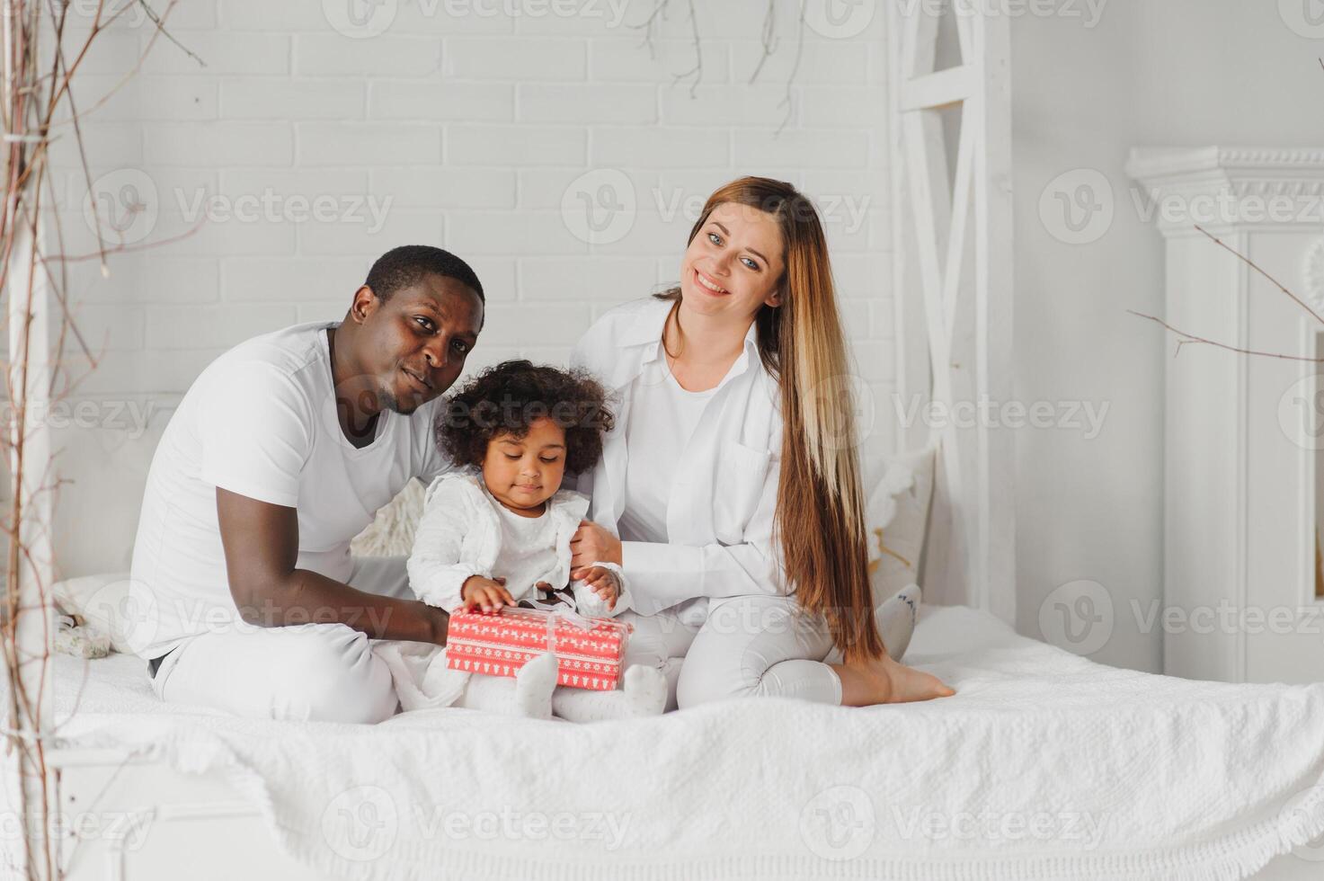 portret van gelukkig multiraciaal jong familie aan het liegen Aan knus wit bed Bij huis, glimlachen Internationale mam en vader ontspannende met weinig biraciaal meisje kind poseren voor afbeelding in slaapkamer foto