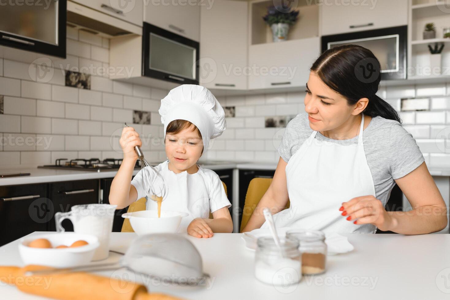 een jong en mooi moeder is voorbereidingen treffen voedsel Bij huis in de keuken, langs met haar weinig zoon foto