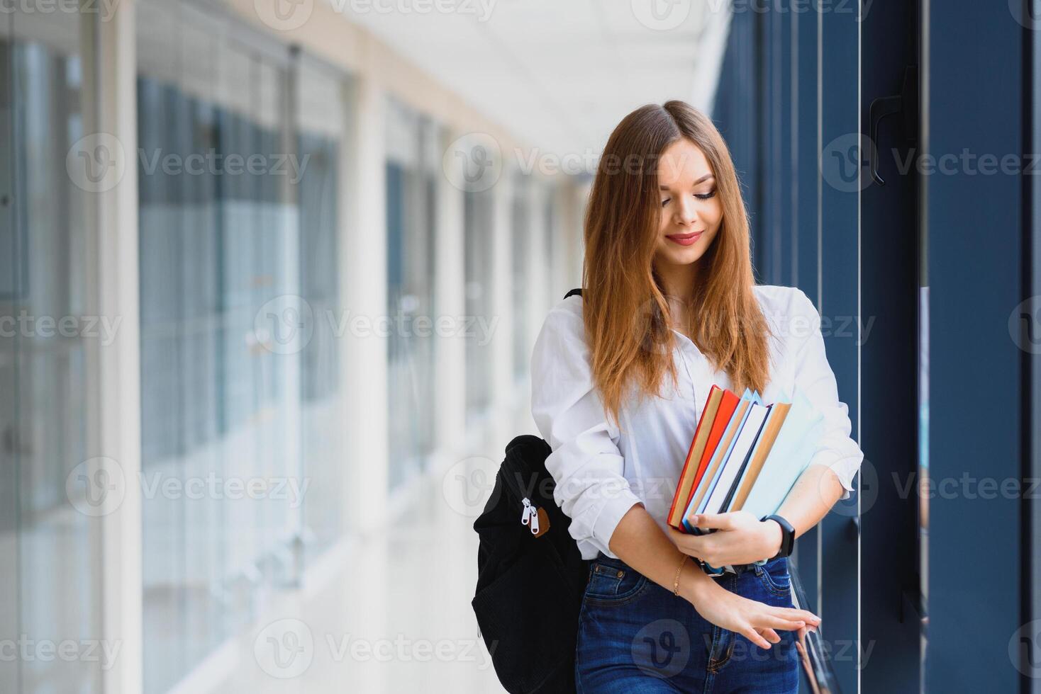 vrolijk brunette leerling meisje met zwart rugzak houdt boeken in modern gebouw. vrouw leerling staand met boeken in college gang foto