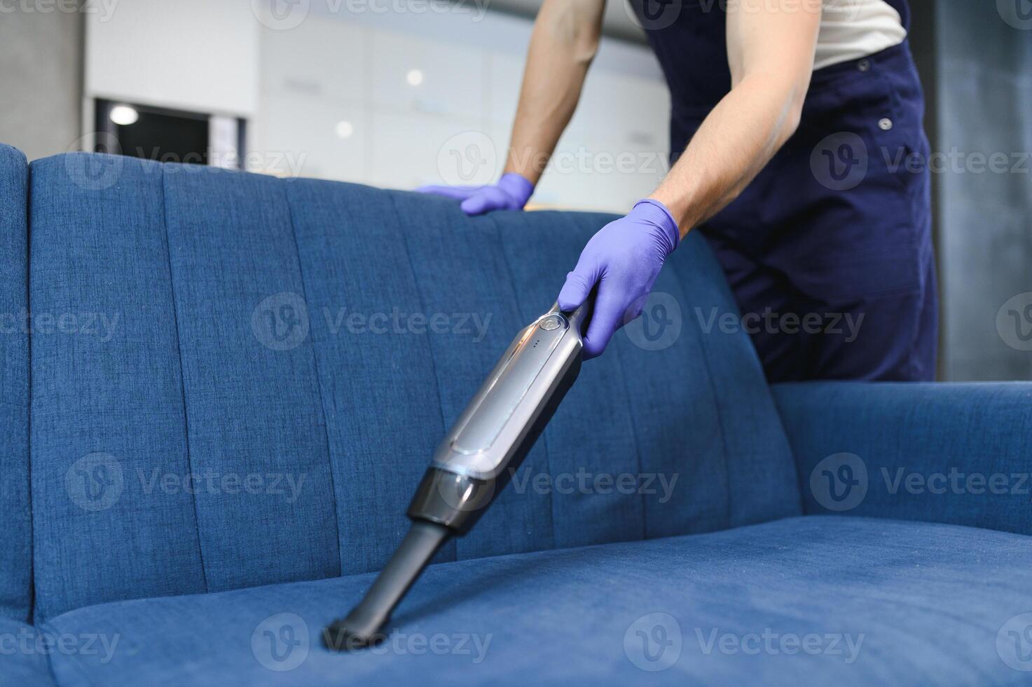 schoonmaak onderhoud. Mens conciërge in handschoenen en uniform vacuüm schoon sofa met professioneel apparatuur. foto