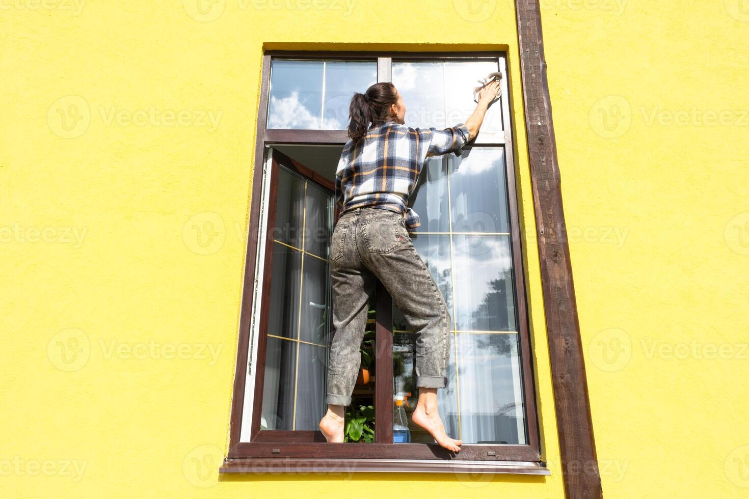 een vrouw handmatig wast de venster van de huis met een vod met een verstuiven schoonmaakster en een dweilen buiten. veiligheid Bij hoogte, herstellen bestellen en netheid in de lente, schoonmaak onderhoud foto