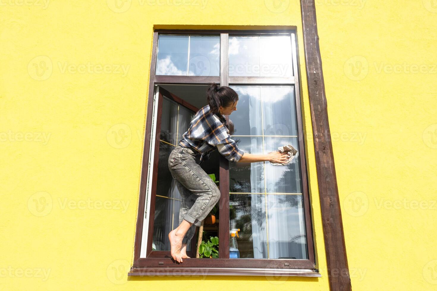 een vrouw handmatig wast de venster van de huis met een vod met een verstuiven schoonmaakster en een dweilen buiten. veiligheid Bij hoogte, herstellen bestellen en netheid in de lente, schoonmaak onderhoud foto