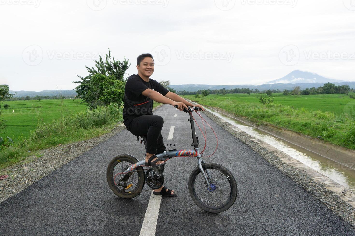 gelukkig Aziatisch Mens rijden een fiets Bij de ochtend- Aan de asfalt weg. wielersport met berg en rijstveld rijst- veld- visie Bij de achtergrond. foto