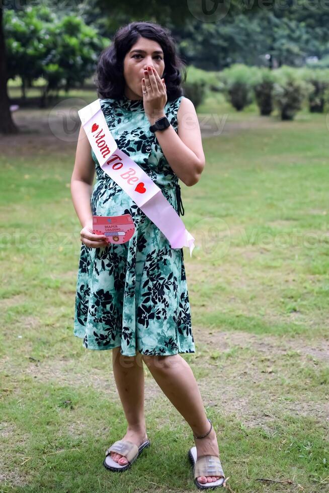 een zwanger Indisch dame poses voor buitenshuis zwangerschap schieten en handen Aan buik, Indisch zwanger vrouw zet haar hand- Aan haar maag met een moederschap jurk Bij maatschappij park, zwanger buiten moederschap schieten foto