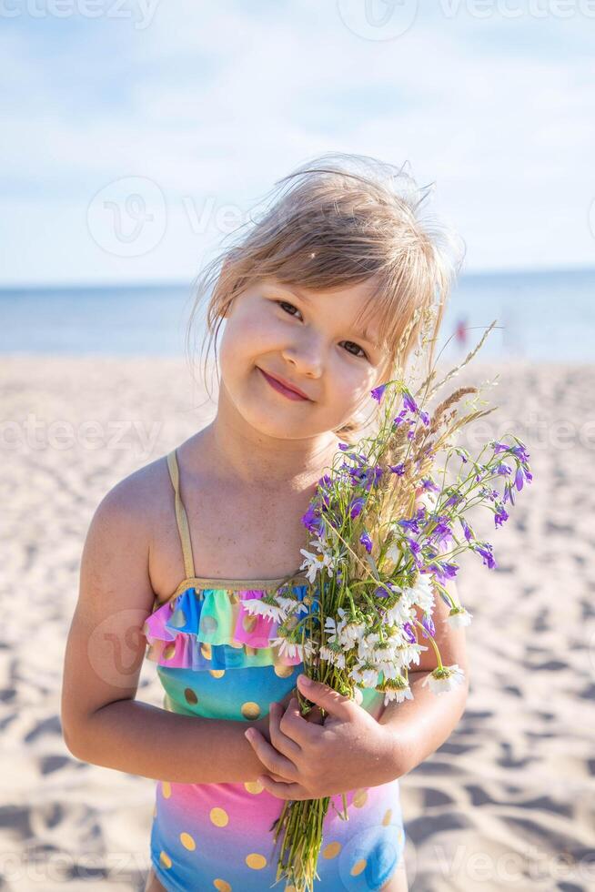 jong gelukkig kind meisje van Europese uiterlijk leeftijd van 6 met bloemen is glimlachen Aan de strand Bij zonnig zomer dag, tropisch zomer roepingen,vakanties.a kind geniet de zee.familie vakantie concept.verticaal foto. foto