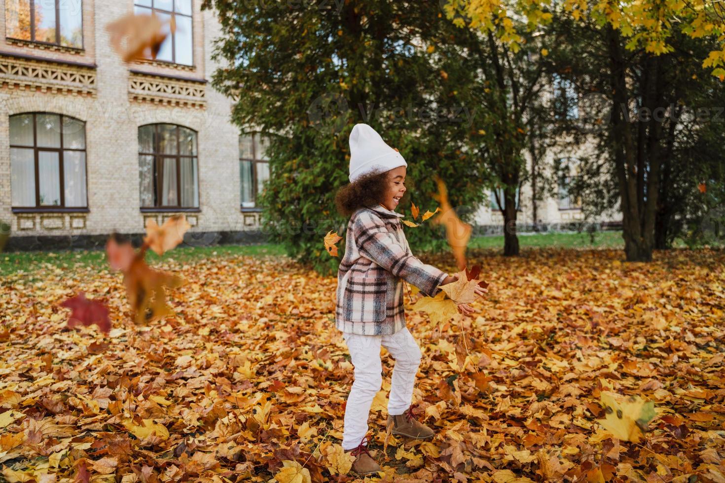zwart meisje met een jas die plezier maakt met gevallen bladeren in het herfstpark foto
