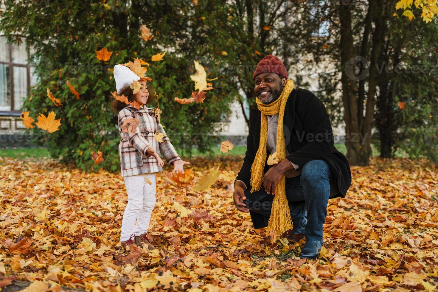 zwarte grootvader en kleindochter maken plezier met gevallen bladeren in het herfstpark foto